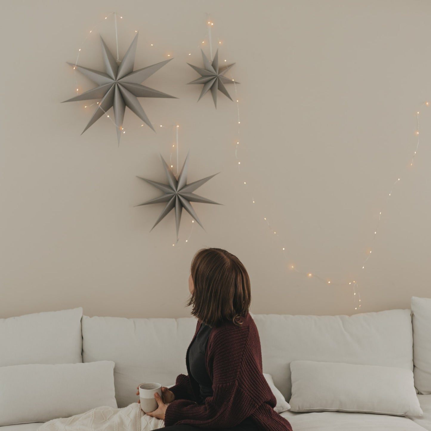 Graue Papiersterne mit warmer Lichterkette als stilvolle Weihnachtsdeko an heller Wand