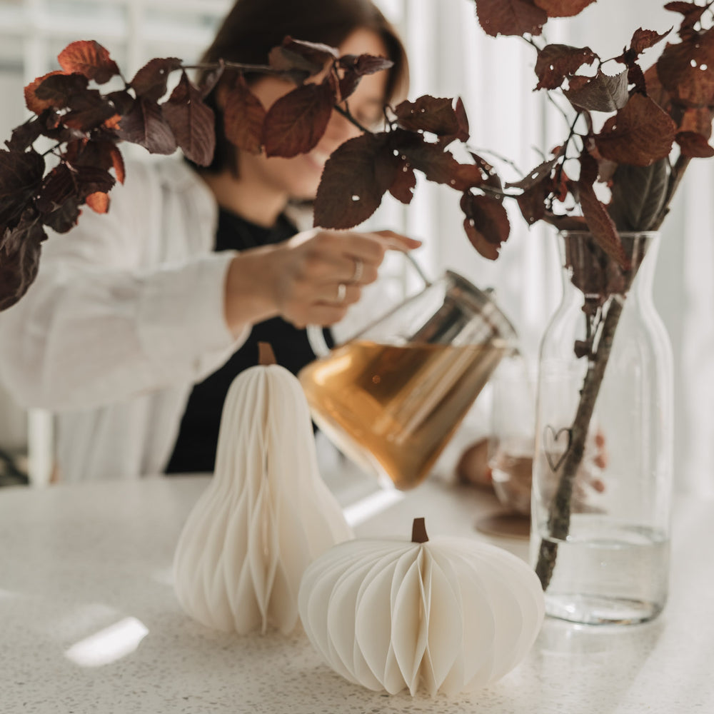 Zwei weiße Papierkürbisse mit Wabenstruktur neben herbstlichem Zweig und Glasvase auf dem Tisch