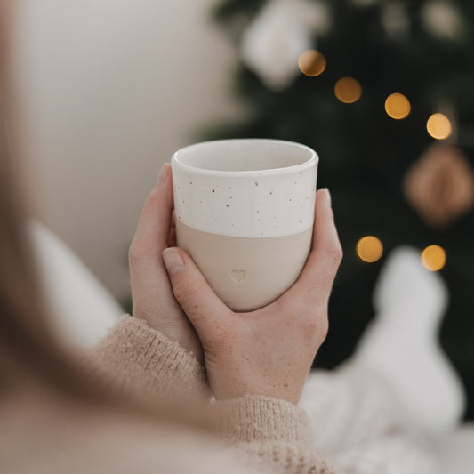 Beigefarbener Keramikbecher von Eulenschnitt mit weiß gesprenkelter Glasur und kleinem Herz, liebevoll in beiden Händen gehalten. Im Hintergrund leuchtet ein geschmückter Weihnachtsbaum mit warmen Lichtern.