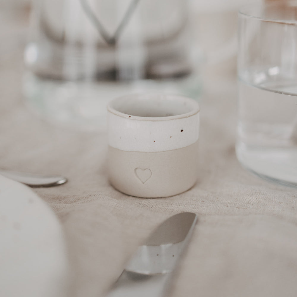 Eierbecher aus handgemachtem Steingut mit Herzprägung auf schlicht gedecktem Tisch neben Glas und Besteck