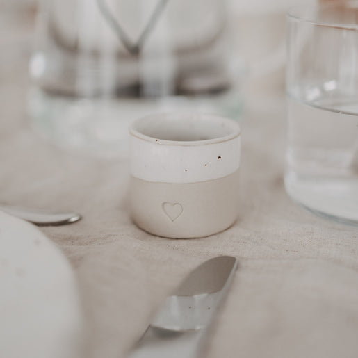 Eierbecher aus handgemachtem Steingut mit Herzprägung auf schlicht gedecktem Tisch neben Glas und Besteck
