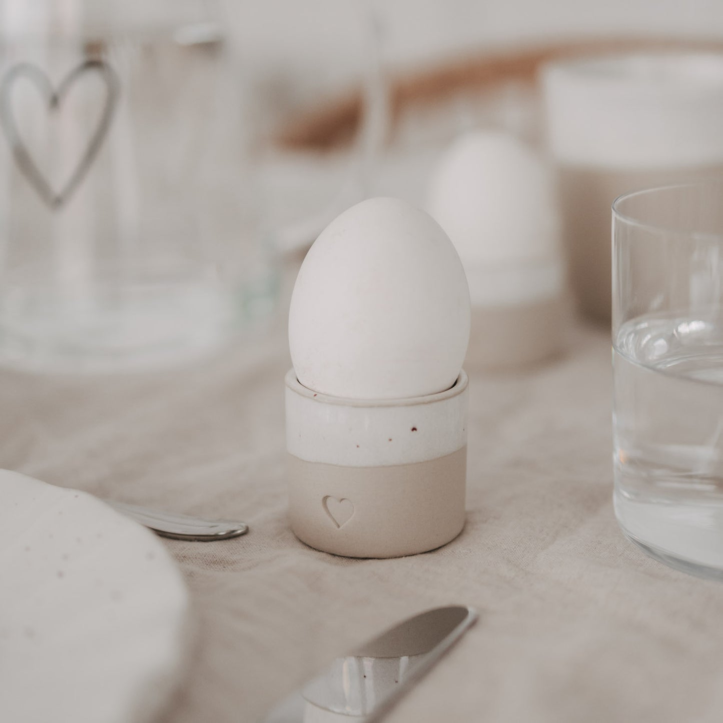 Eierbecher aus handgemachtem Steingut mit Herzprägung und weißem Ei auf gedecktem Frühstückstisch