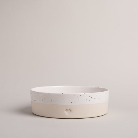 Flache, zweifarbige Keramikschale in Beige und Weiß mit feinen Sprenkeln. Auf der Vorderseite befindet sich ein eingestanztes Herzsymbol. Die Schale steht auf hellem Untergrund vor neutralem Hintergrund.
