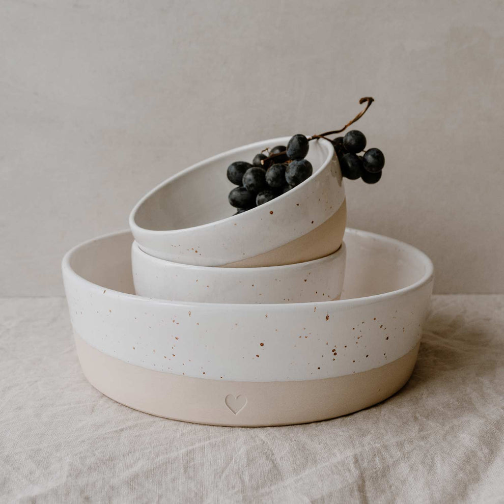 Drei ineinander gestapelte Keramikschalen in verschiedenen Größen, alle mit zweifarbiger Gestaltung in Beige und Weiß, feiner Sprenkelung und eingestanztem Herz. In der obersten Schale liegt eine Traube dunkler Weintrauben. Die Schalen stehen auf einem hellen Leinentuch.