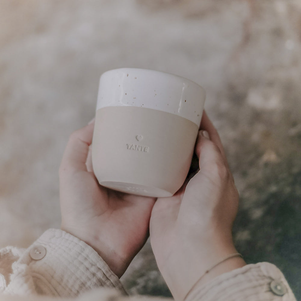 Zwei Hände halten einen minimalistischen Keramikbecher in sanftem Beige mit cremeweißer, gesprenkelter Glasur am oberen Rand. Auf der Vorderseite ist ein kleines Herz und darunter das Wort ‚TANTE‘ eingeprägt. Der Becher wird leicht schräg vor einem natürlichen, rauen Hintergrund präsentiert – ein stilvolles Geschenk mit persönlicher Botschaft.