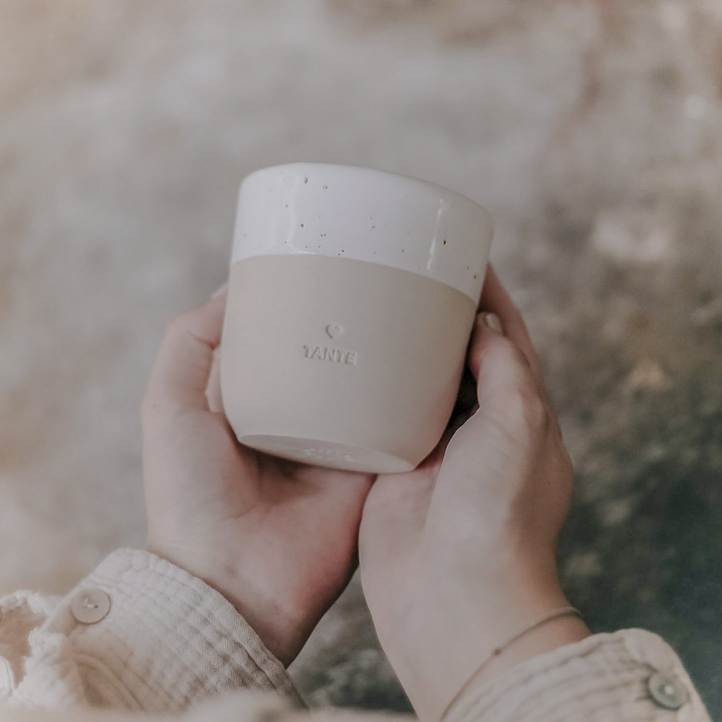 Zwei Hände halten einen minimalistischen Keramikbecher in sanftem Beige mit cremeweißer, gesprenkelter Glasur am oberen Rand. Auf der Vorderseite ist ein kleines Herz und darunter das Wort ‚TANTE‘ eingeprägt. Der Becher wird leicht schräg vor einem natürlichen, rauen Hintergrund präsentiert – ein stilvolles Geschenk mit persönlicher Botschaft.
