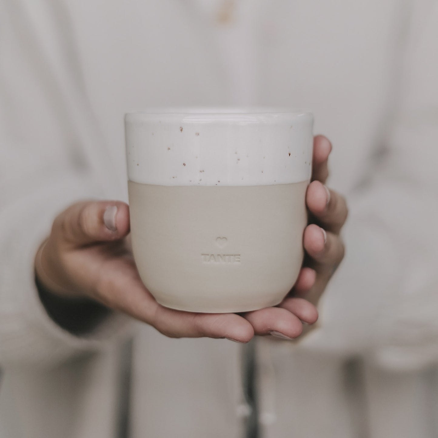 Hand hält einen schlichten, beigen Keramikbecher mit cremeweißer Glasur am oberen Rand. Auf der Vorderseite ist ein kleines Herz und darunter das Wort ‚TANTE‘ eingeprägt. Der Becher wird in beiden Händen vor dem Körper präsentiert – stilvoll, minimalistisch und mit liebevoller Botschaft, ideal als Geschenk für die Tante.