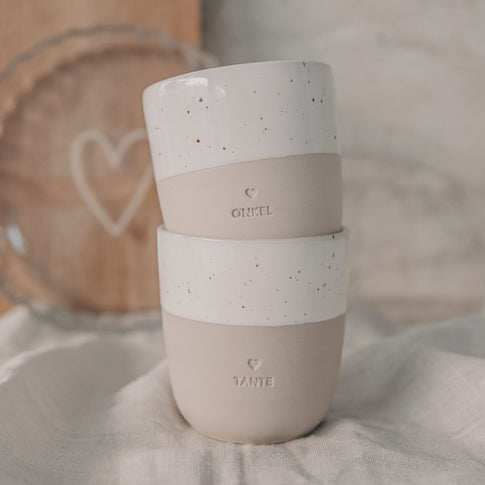 Keramikbecher ONKEL in Beige-Weiß mit Speckle-Glasur, wird in beiden Händen gehalten.