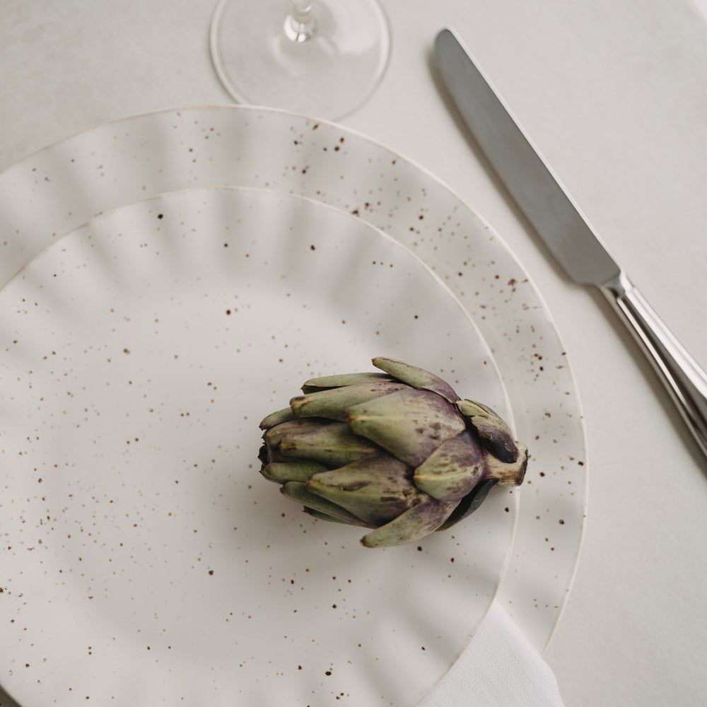 Zwei Colina-Teller übereinander auf weißer Tischdecke mit Artischocke als Deko