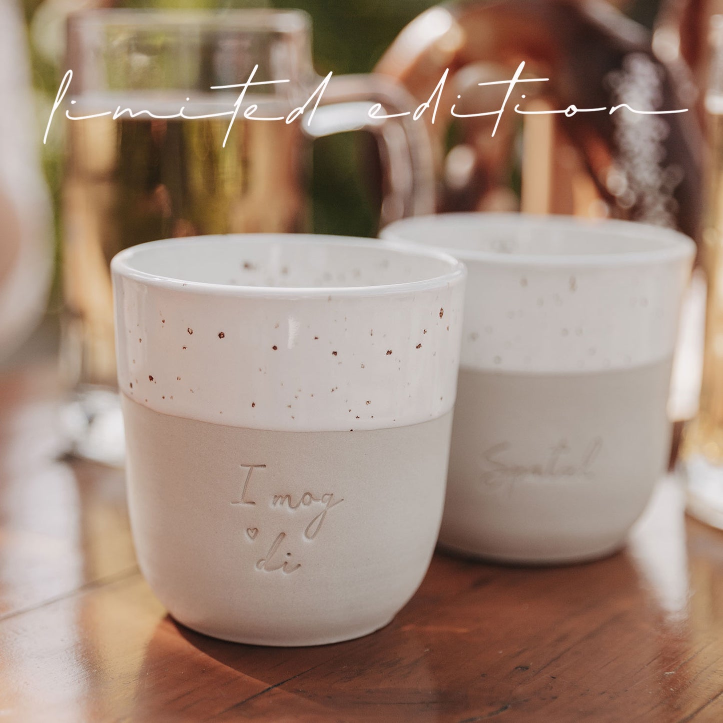 Detailansicht der Becher „I mog di“ und „Spatzl“ mit Bierkrügen im Hintergrund im Biergarten.