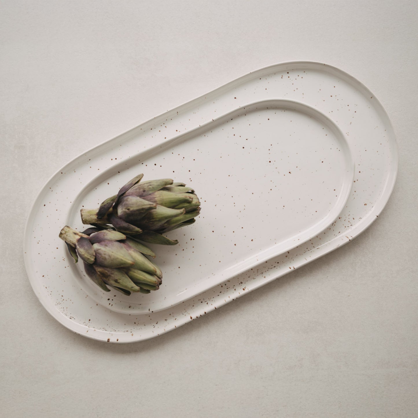 Servierplatten Calma in zwei Größen auf hellem Tisch – Detail mit Artischocken als Deko