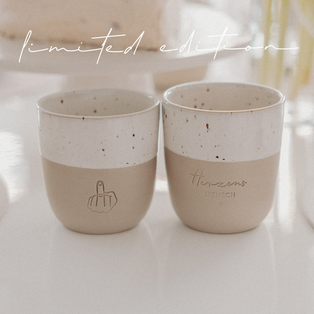 Zwei Limited-Edition-Becher in Beige und Weiß mit Herzensmensch- und Mittelfinger-Motiv auf hellem Tisch arrangiert.