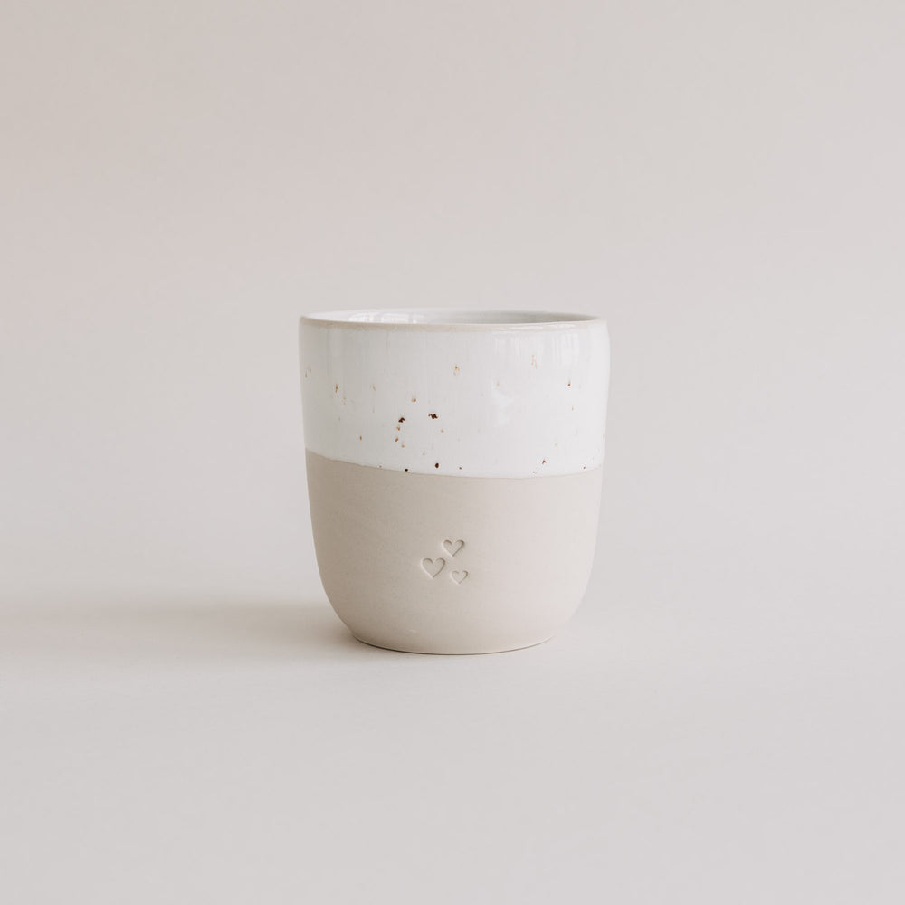 Keramikbecher in Beige und Weiß mit drei Herzprägungen, minimalistisch fotografiert vor hellem Hintergrund.