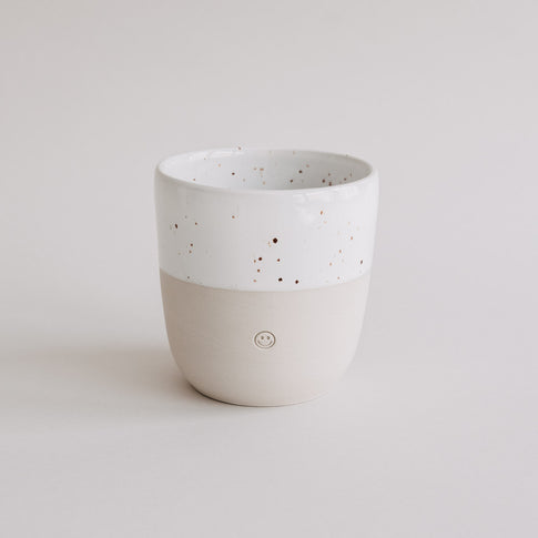 Heller Keramikbecher mit weißer Sprenkelglasur und eingeprägtem Smiley, freigestellt vor neutralem Hintergrund.