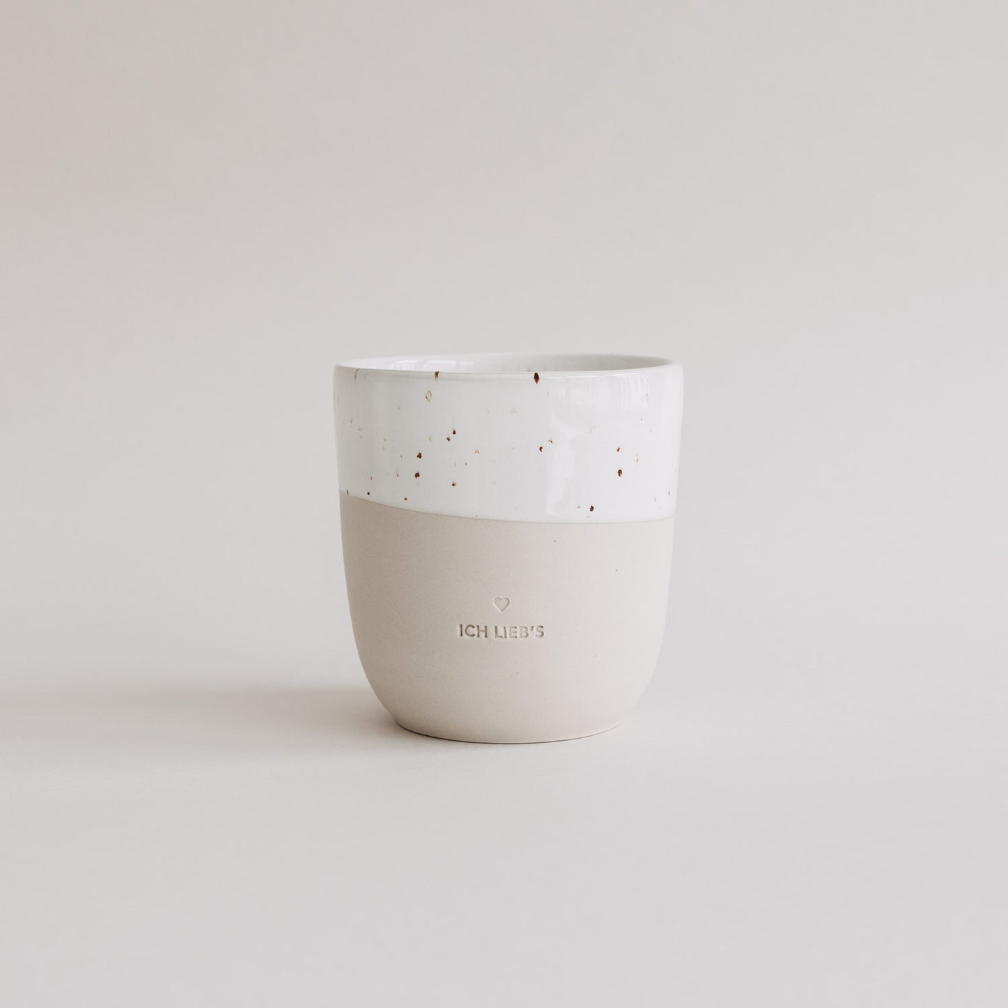 Keramiktasse in Beige und Weiß mit Sprenkeln und Prägung „Ich lieb’s“ vor hellem Hintergrund.