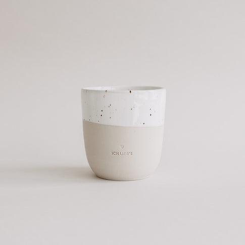 Keramiktasse in Beige und Weiß mit Sprenkeln und Prägung „Ich lieb’s“ vor hellem Hintergrund.