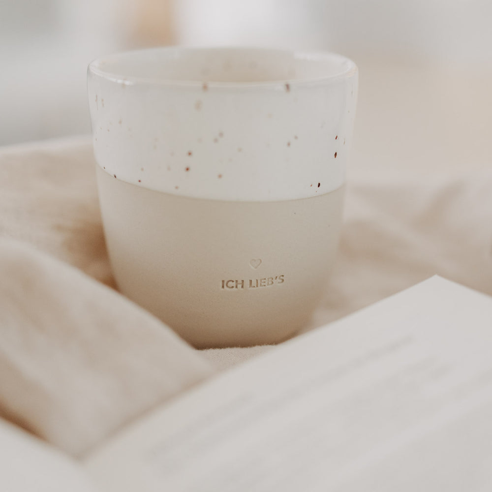 Beige-weiße „Ich lieb’s“-Tasse mit Sprenkelglasur auf Stoff, neben geöffnetem Buch.