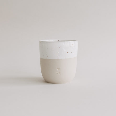 Heller Keramikbecher mit weißer Sprenkelglasur und MR-Prägung auf beigefarbener Unterseite vor neutralem Hintergrund.