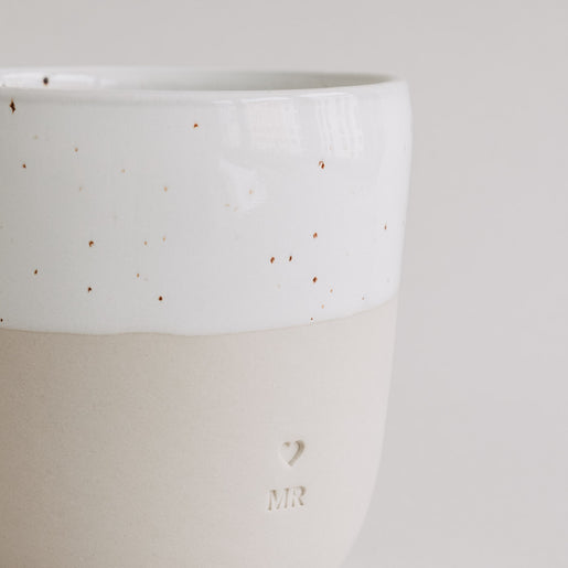 Seitliche Nahaufnahme des MR-Bechers mit Herz- und MR-Prägung auf matter beiger Keramik.