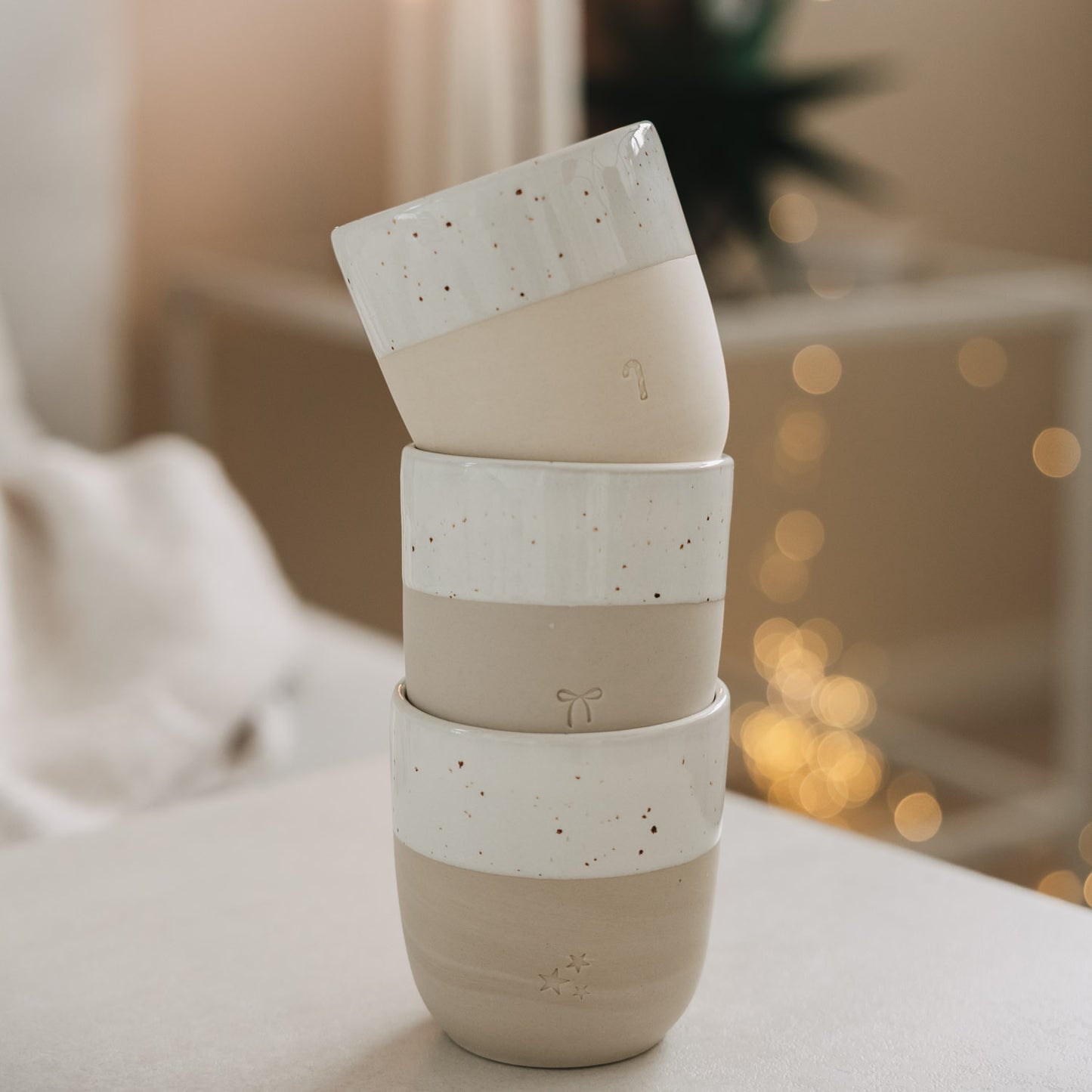 Gestapelte Keramikbecher mit Schleifen- und Sternmotiv, minimalistische Weihnachtsdeko mit Lichterhintergrund
