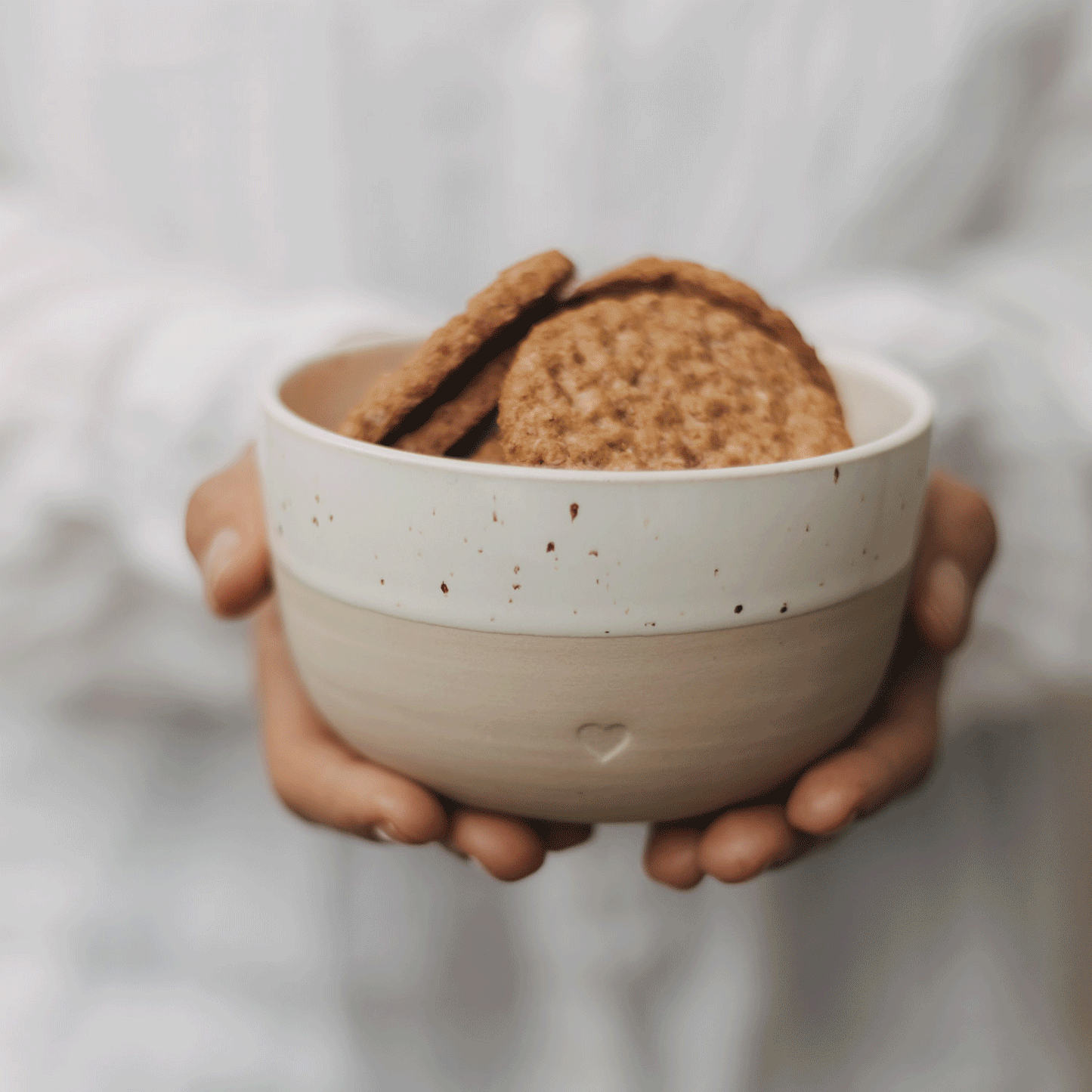 Keramikschale mit Herz in Beige und Weiß, gefüllt mit Keksen und in den Händen gehalten