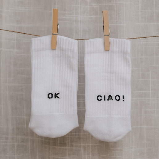 Weiße Socken mit schwarzem Text ‚OK‘ auf der einen und ‚CIAO!‘ auf der anderen Ferse, hängen an einer Wäscheleine.