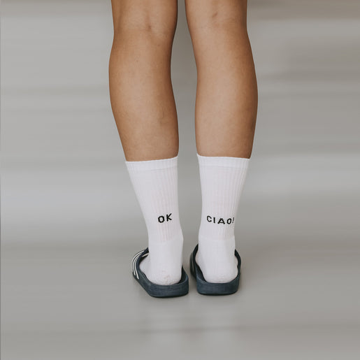 Nahaufnahme: Frau trägt weiße Socken mit schwarzem Schriftzug ‚OK‘ und ‚CIAO!‘ auf den Fersen und trägt Latschen.
