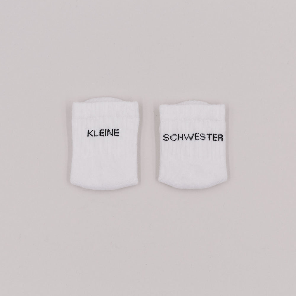Zwei weiße Babysocken mit schwarzem Schriftzug „Kleine Schwester“ auf hellem Hintergrund.
