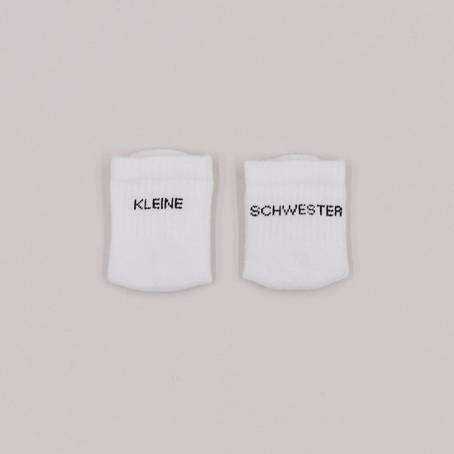Zwei weiße Babysocken mit schwarzem Schriftzug „Kleine Schwester“ auf hellem Hintergrund.