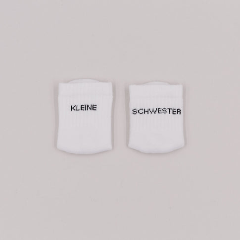 Zwei weiße Babysocken mit schwarzem Schriftzug „Kleine Schwester“ auf hellem Hintergrund.