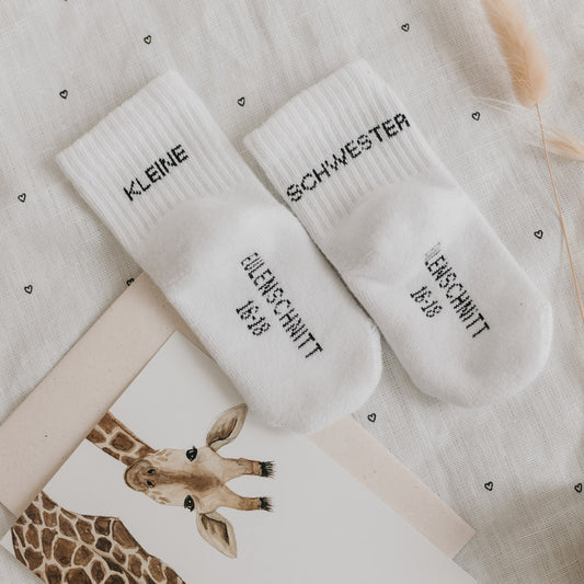 Weiße Babysocken „Kleine Schwester“ mit Markenaufdruck, dekoriert mit Giraffenkarte und Trockenblumen.