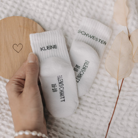 Hand hält Babysocken „Kleine Schwester“ auf strukturierter Decke mit Holzuntersetzer und Blättern.