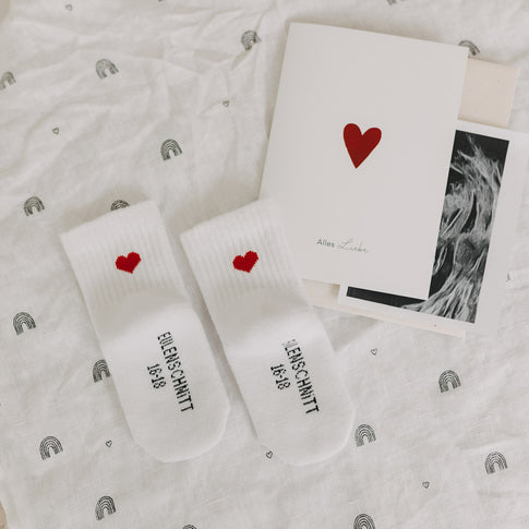 Babysocken mit Herzmotiv auf Stoff mit Regenbogenmuster, daneben Karte mit Herz und Ultraschallfoto – Geschenk zur Schwangerschaft.