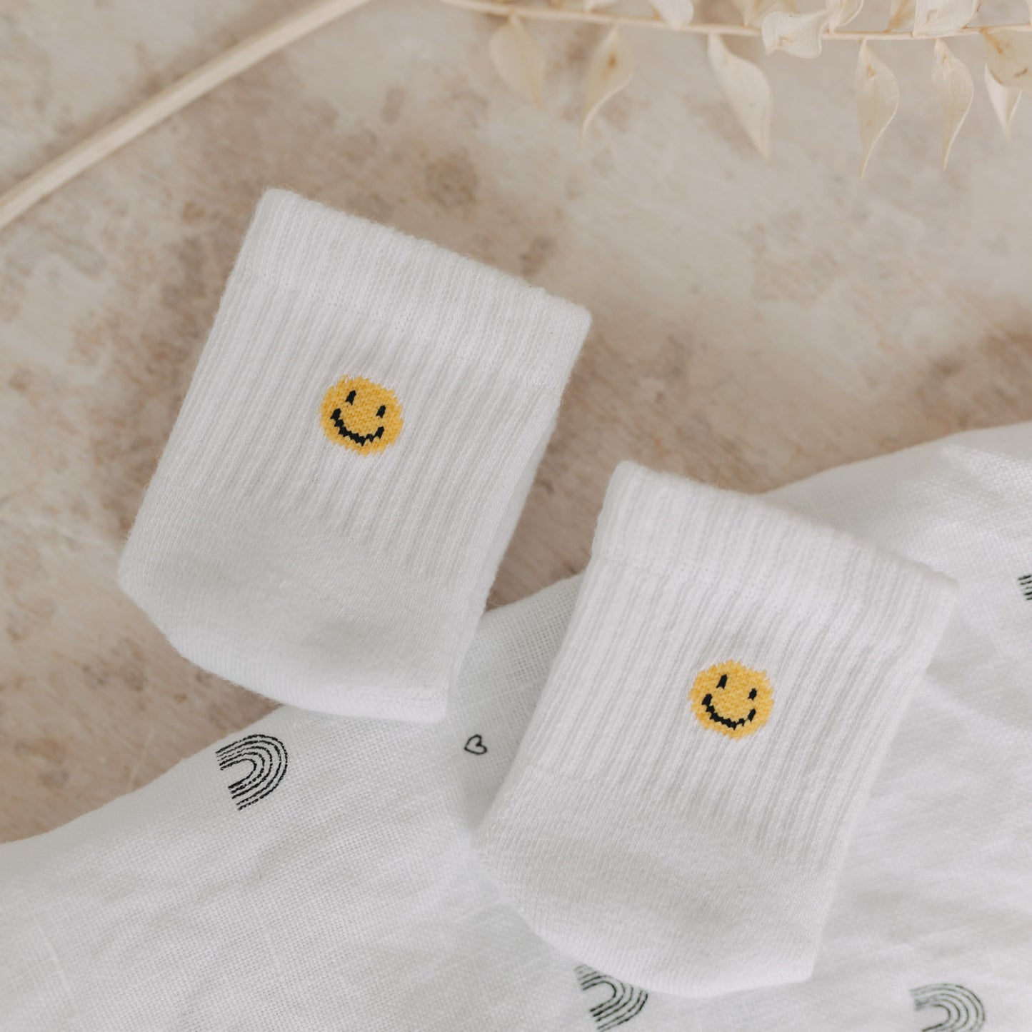 Weiße Babysocken mit gelbem Smiley auf gemustertem Stoff, dekoriert mit Trockenblumen.