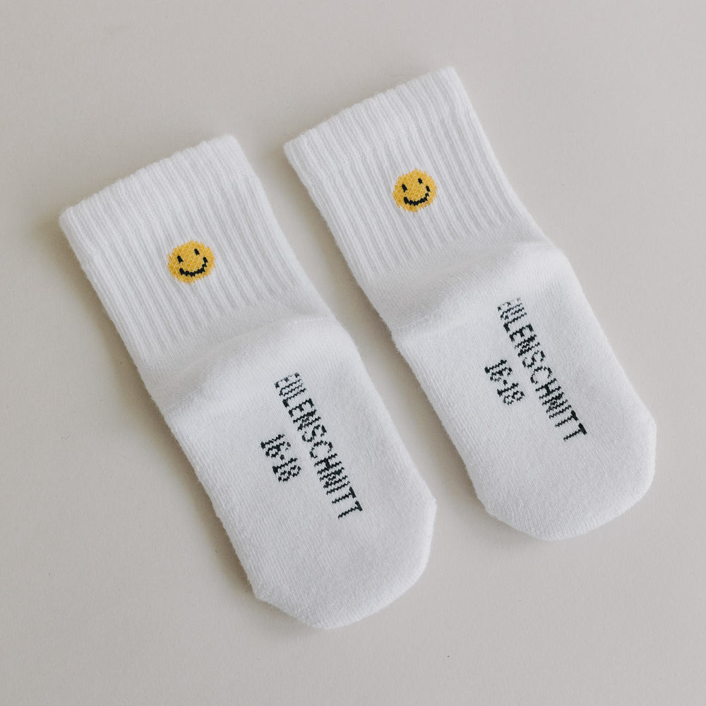Weiße Babysocken mit gelbem Smiley und Eulenschnitt-Schriftzug auf glattem, hellem Hintergrund.
