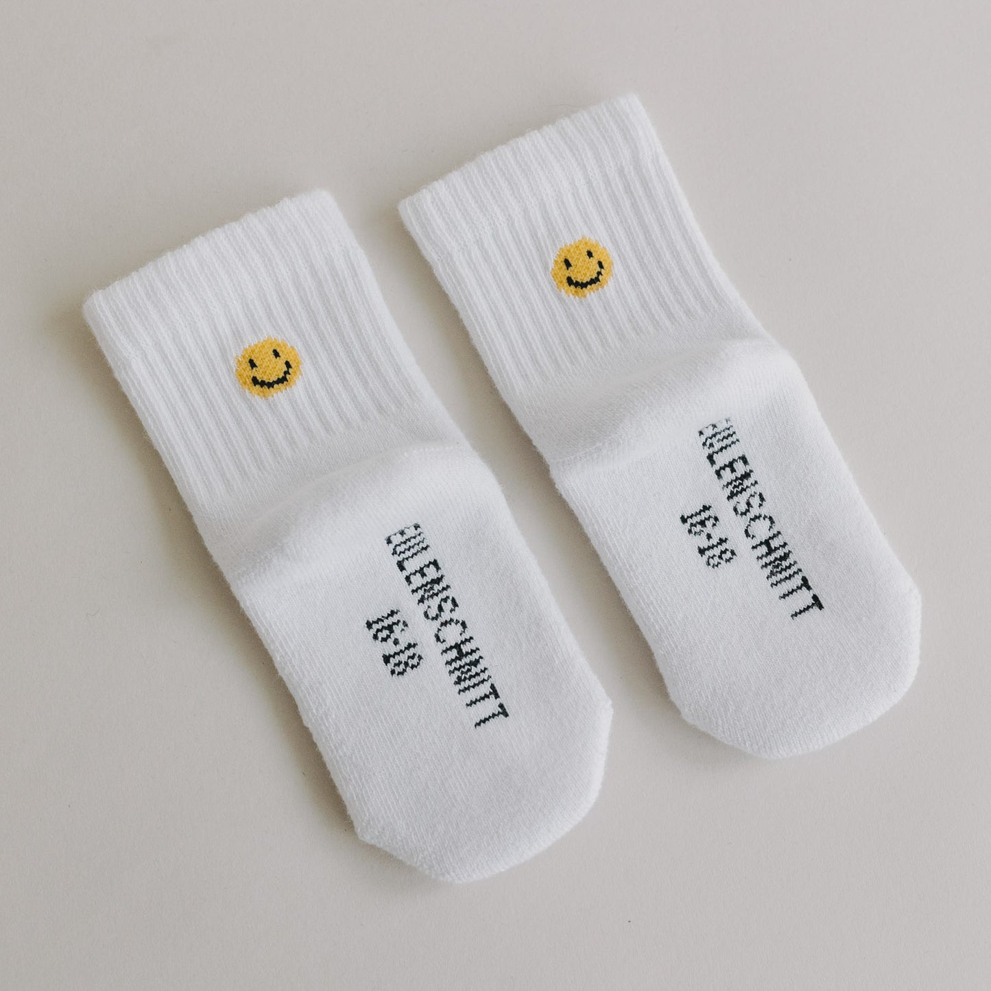 Weiße Babysocken mit gelbem Smiley und Eulenschnitt-Schriftzug auf glattem, hellem Hintergrund.