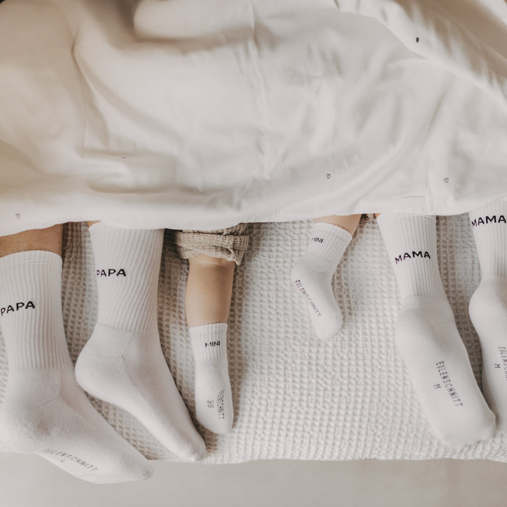 Familienfüße im Bett: Eltern und Kind mit weißen Socken in Mama-, Papa- und Mini-Version unter einer Decke.