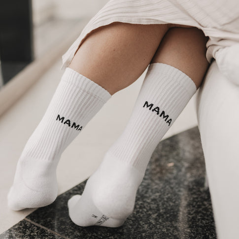 Frau sitzt entspannt mit übergeschlagenen Beinen und trägt weiße Socken mit Mama-Schriftzug.