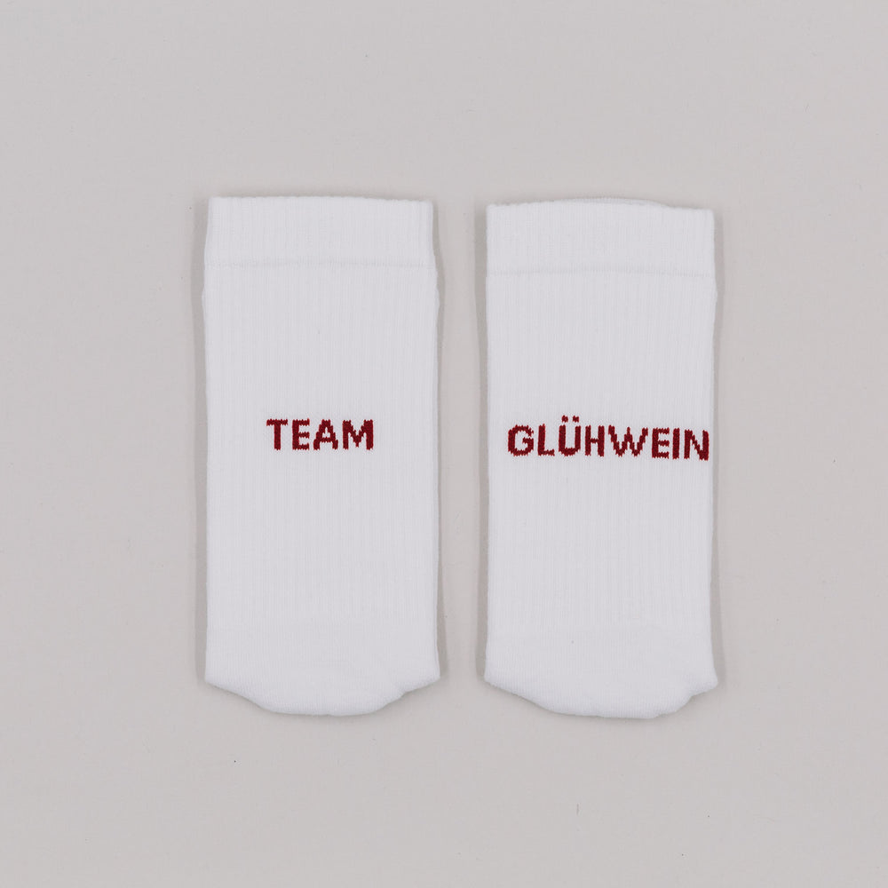 Weiße Socken mit rotem Schriftzug „Team Glühwein“ nebeneinander liegend vor neutralem Hintergrund.
