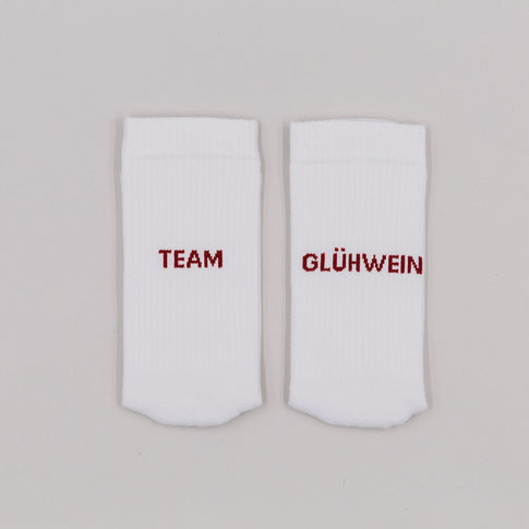 Weiße Socken mit rotem Schriftzug „Team Glühwein“ nebeneinander liegend vor neutralem Hintergrund.