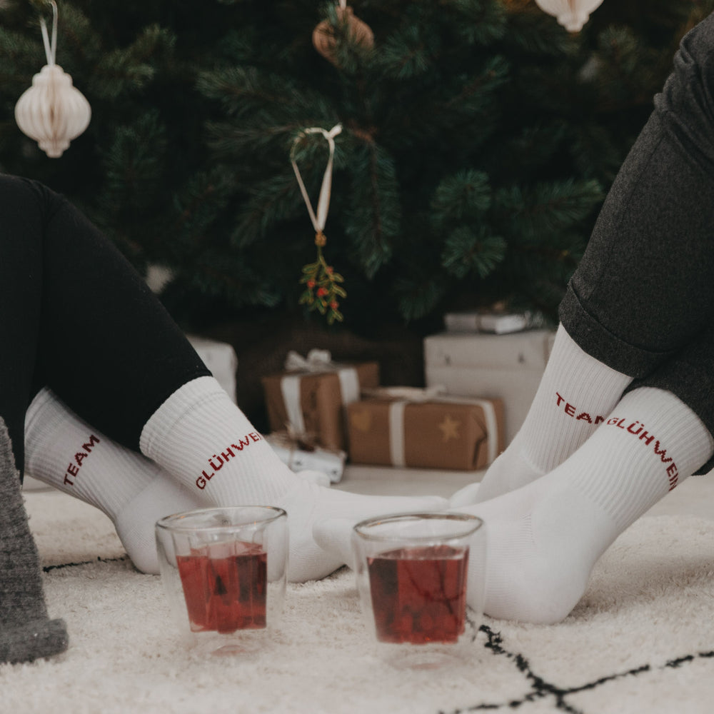 Vier Füße in weißen „Team Glühwein“-Socken vor einem Weihnachtsbaum, zwei Glühweingläser auf dem Teppich.