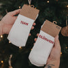 Zwei Hände halten Socken mit rotem Schriftzug „Team Glühwein“ vor einem geschmückten Weihnachtsbaum.