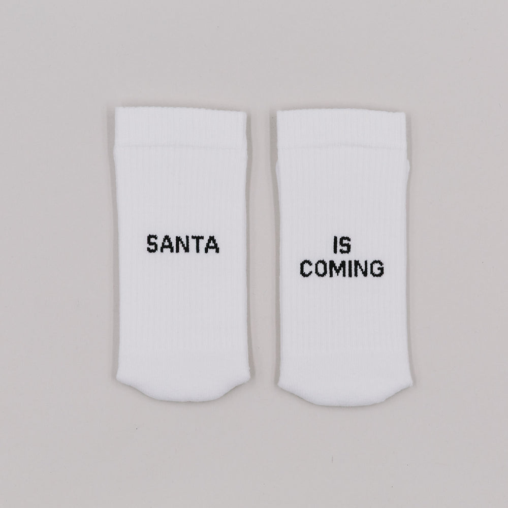 Weiße Socken mit schwarzem Schriftzug „Santa is coming“ nebeneinander auf hellem Hintergrund
