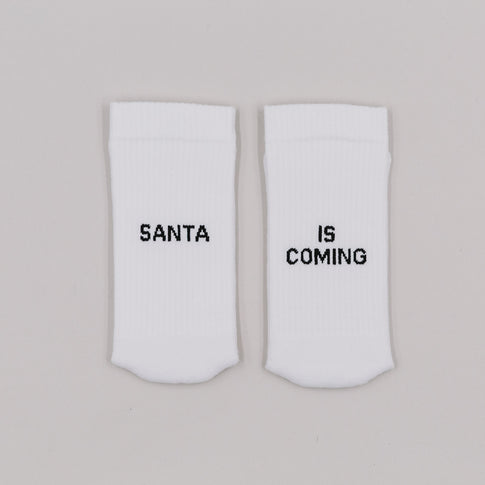 Weiße Socken mit schwarzem Schriftzug „Santa is coming“ nebeneinander auf hellem Hintergrund