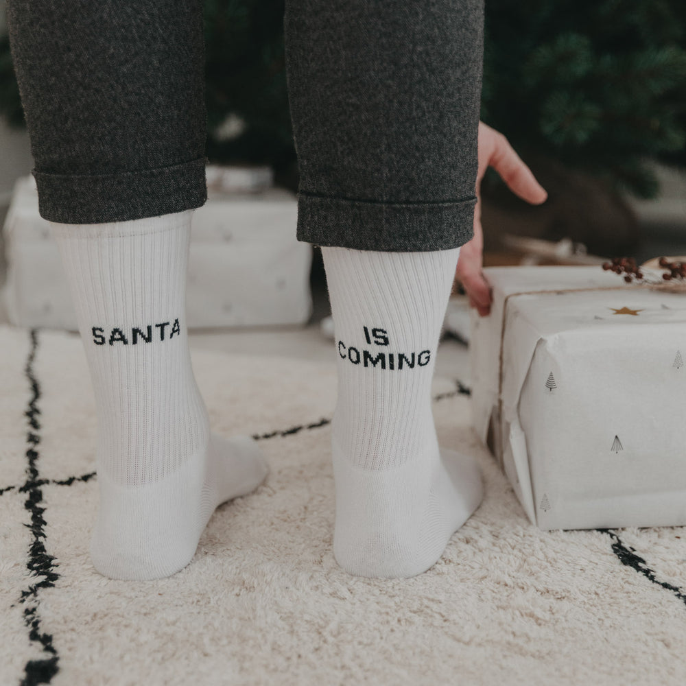 Person mit „Santa is coming“-Socken beugt sich über Geschenk vor dem Weihnachtsbaum