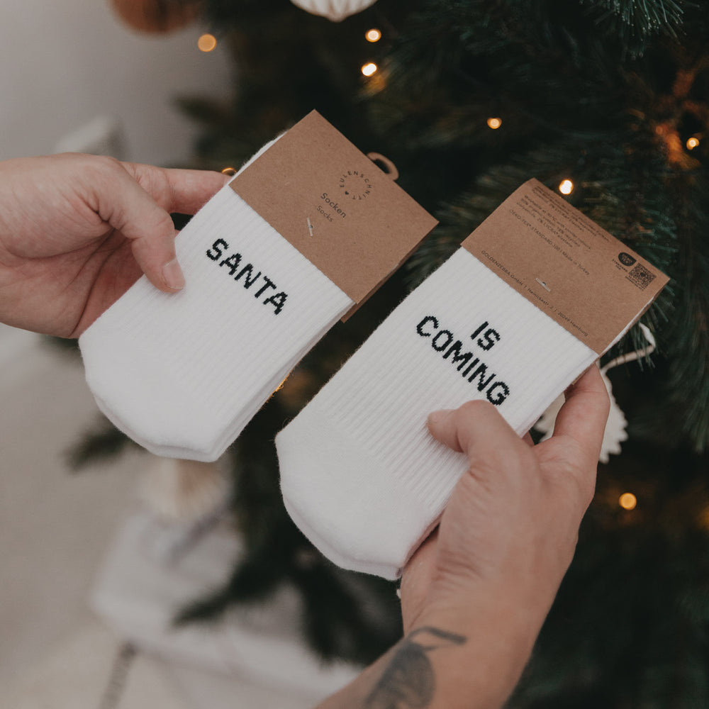 Nahaufnahme der verpackten „Santa is coming“-Socken mit Kartonverpackung vor Weihnachtsbaum