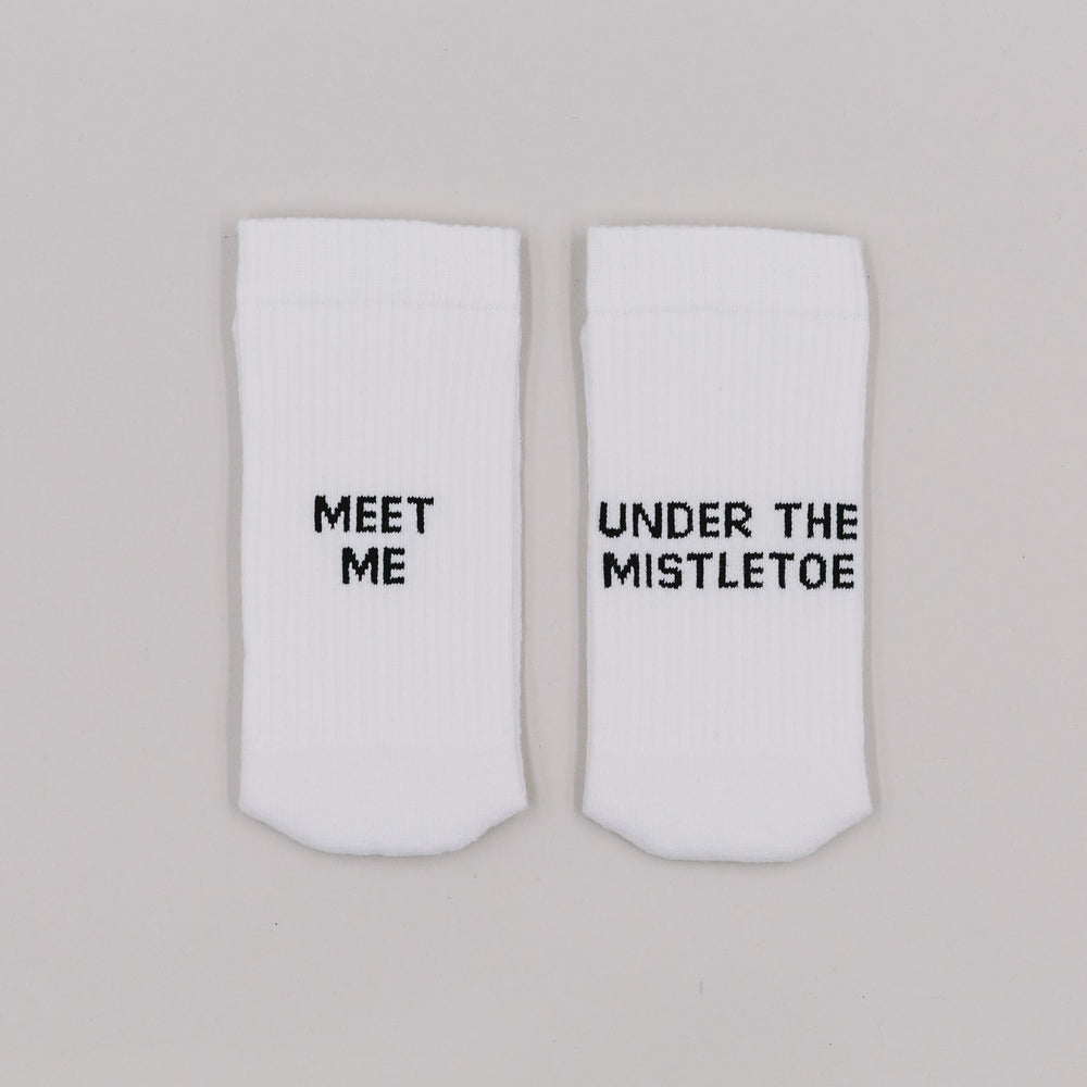Weiße Socken mit der Aufschrift „Meet me“ und „Under the mistletoe“ auf grauem Hintergrund nebeneinander liegend
