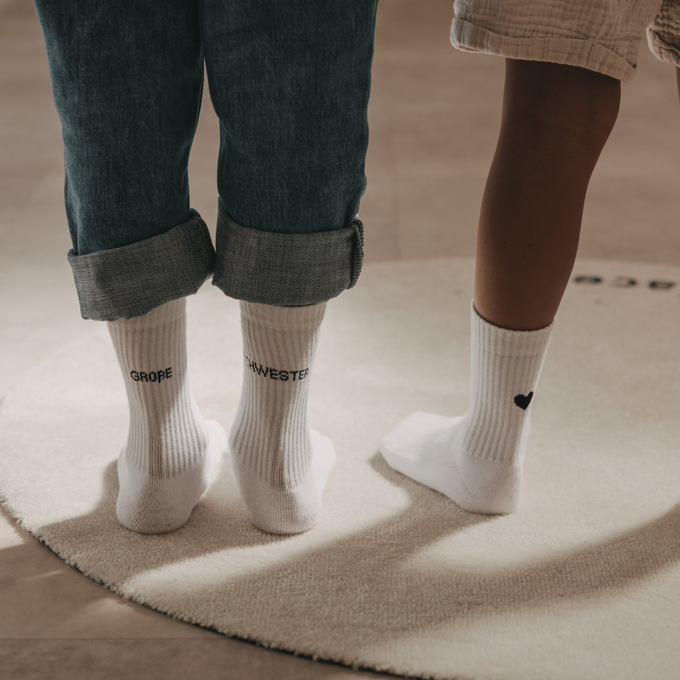 Kids Socken Große Schwester in Weiß mit schwarzem Schriftzug neben weißen Kindersocken mit Herzdetail