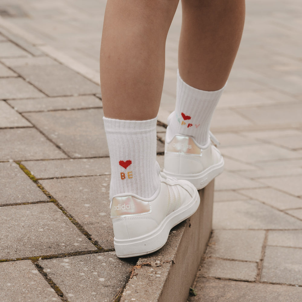Weiße Kinder Socken „Be Happy“ mit Herz, kombiniert mit Sneakern auf Bordsteinkante