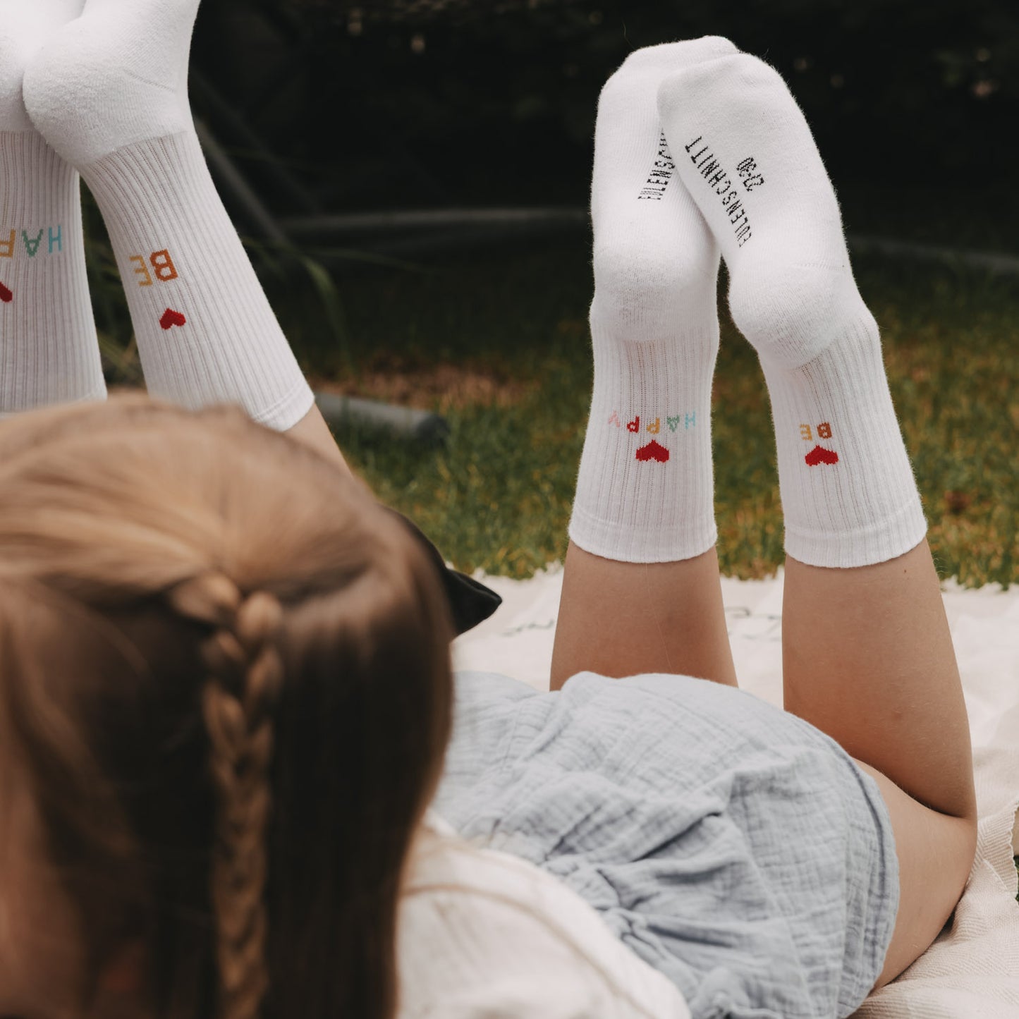 Kinder liegen im Gras und tragen weiße „Be Happy“ Socken mit Herzmotiv